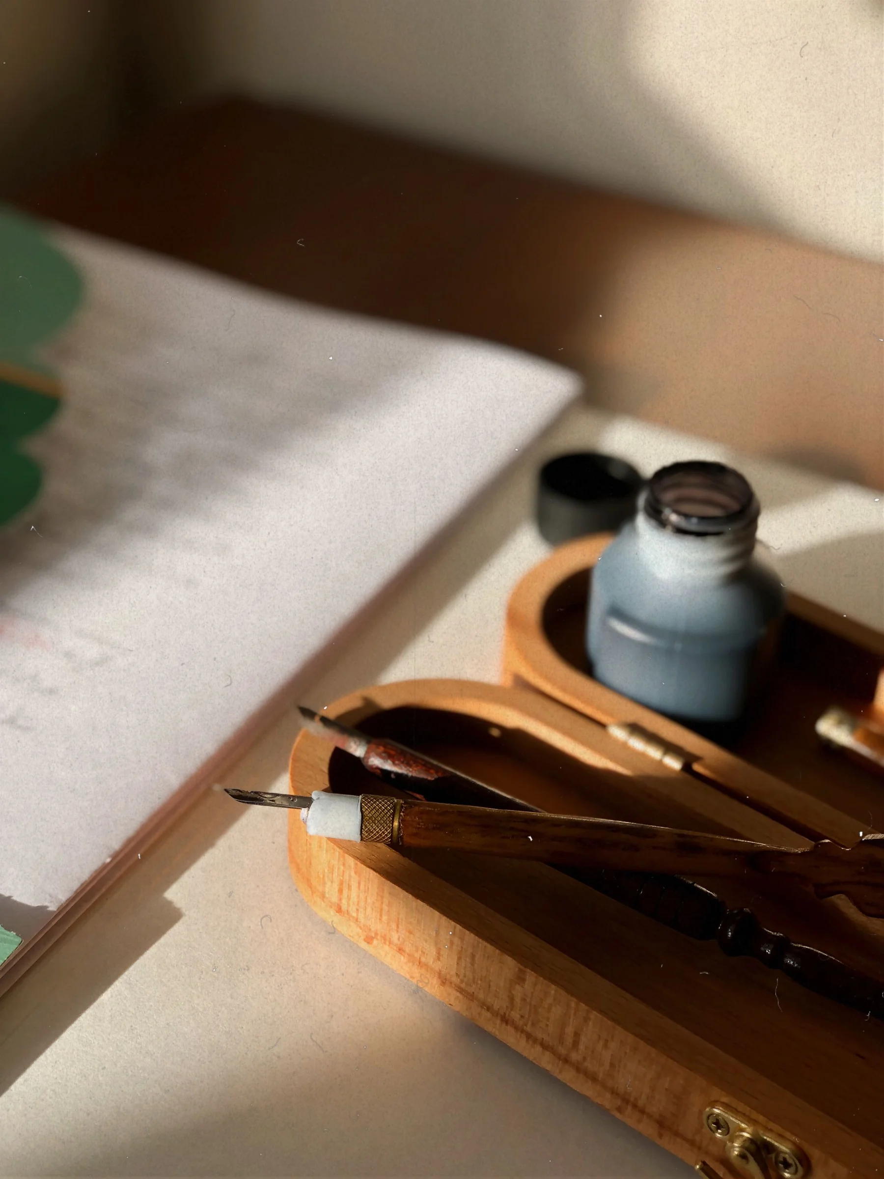 Metal dip-pen nibs rest in a wooden holder beside an ink bottle and an open notebook.