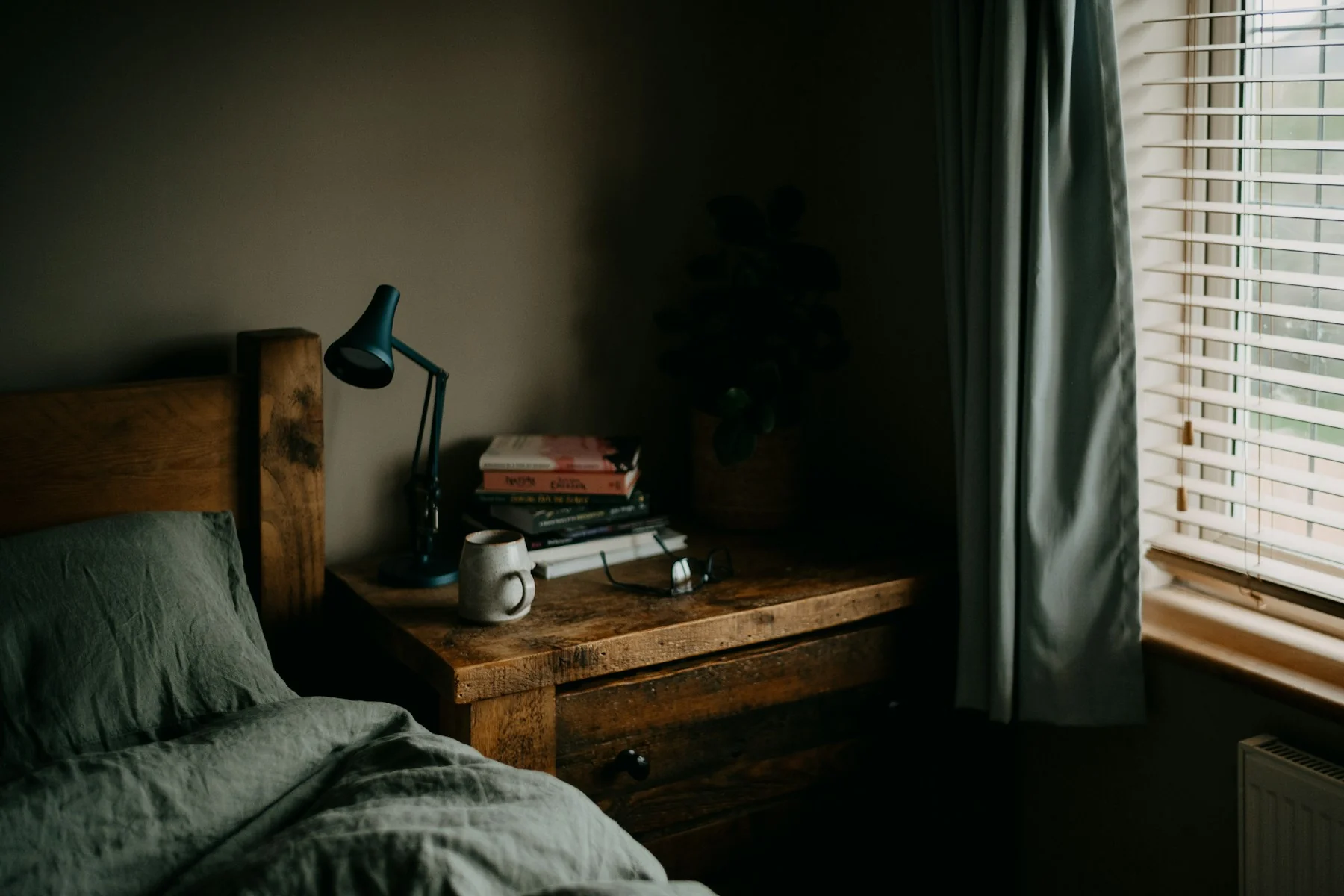 A bedroom with muted green bedding, a wooden bedhead and a reading lamp beside a window.
