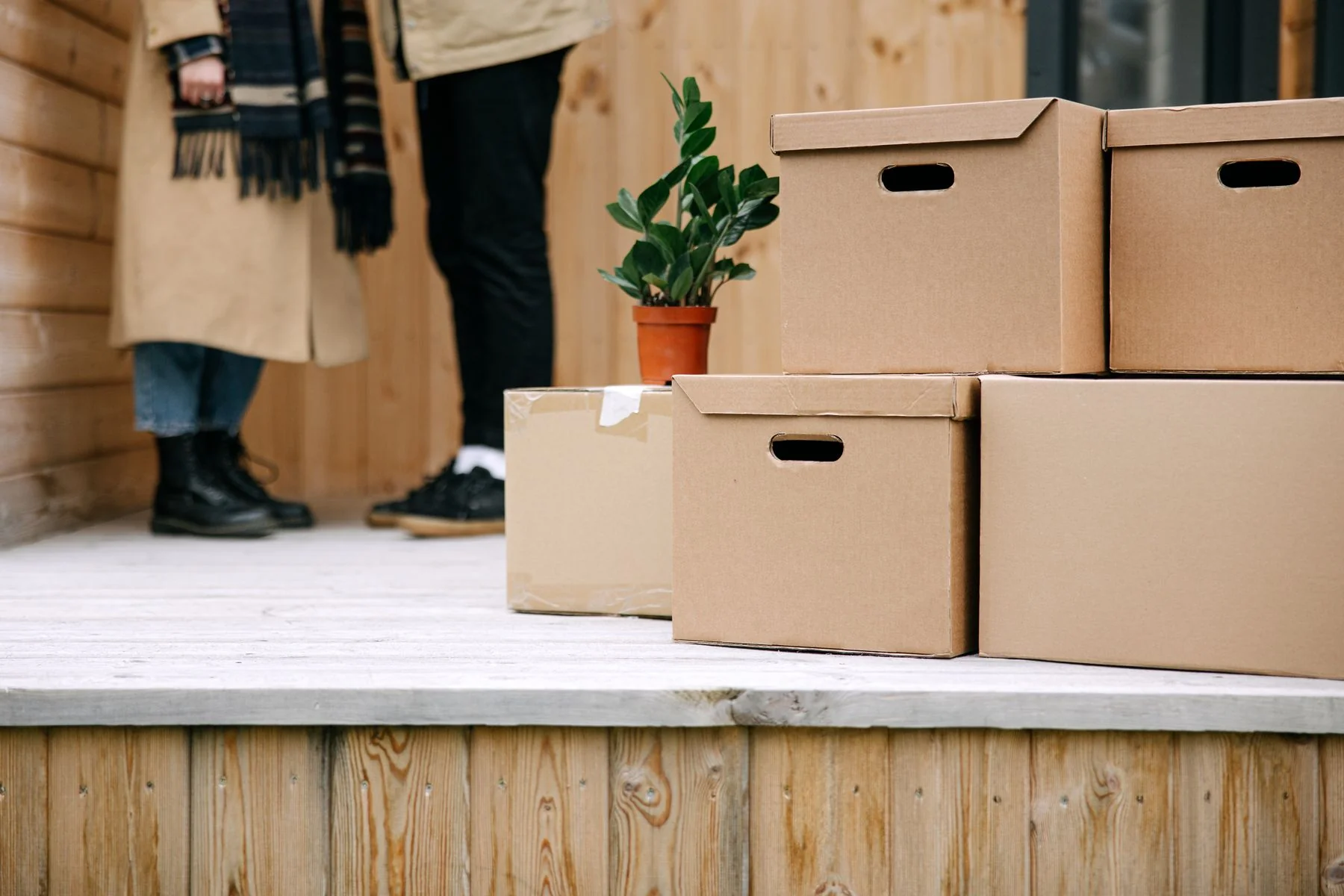 Cardboard moving boxes and a small green plant on a wooden porch, with people in the background.