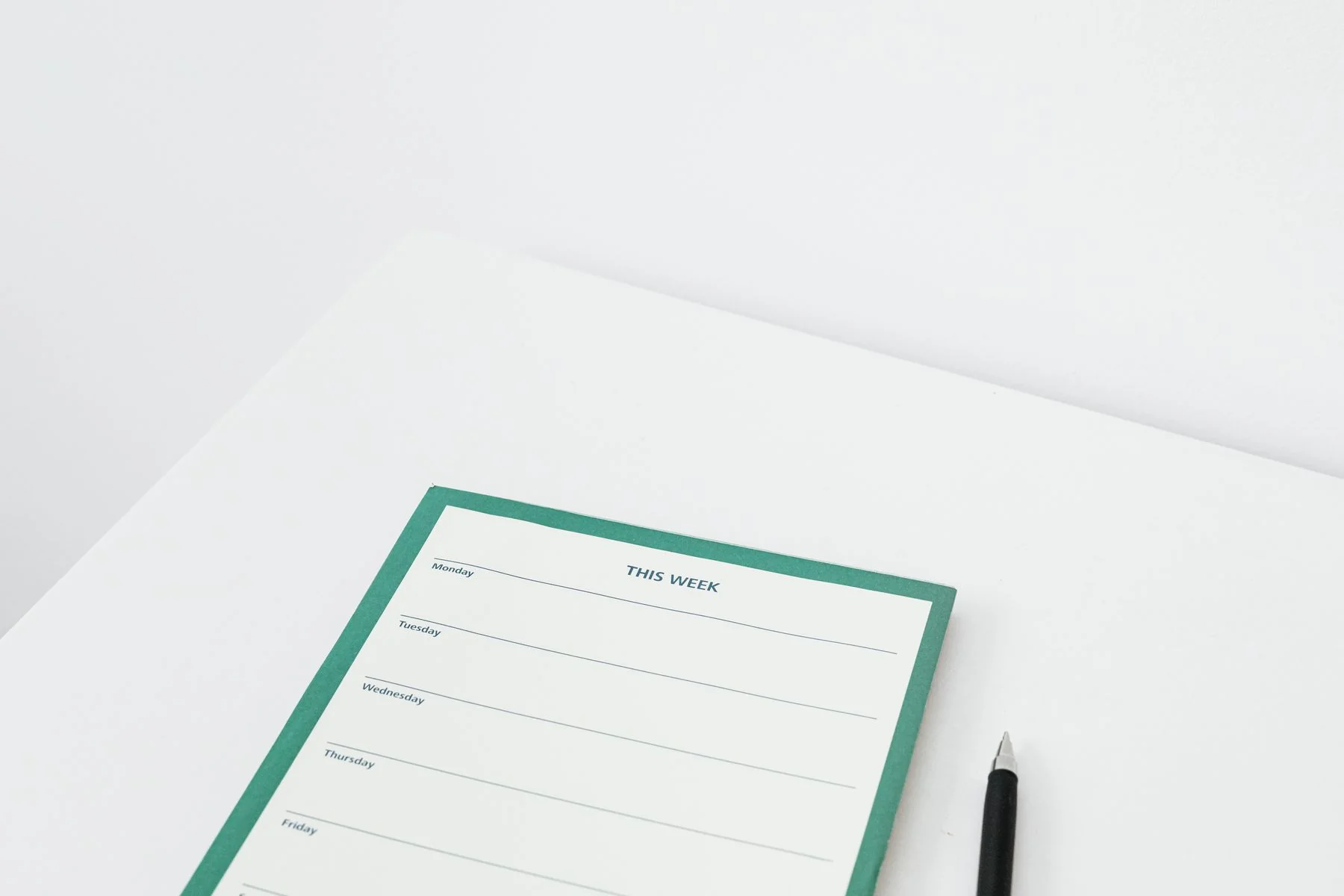 A weekly planner with a muted green border and a black pen on a white desk.