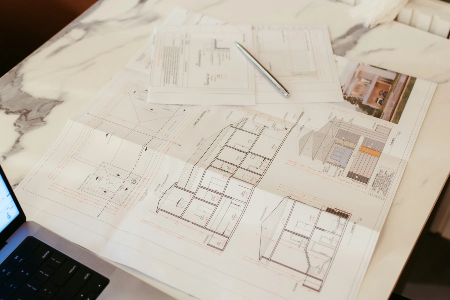 Architectural floor plans and a pencil on a pale stone desk, with a laptop at the edge.