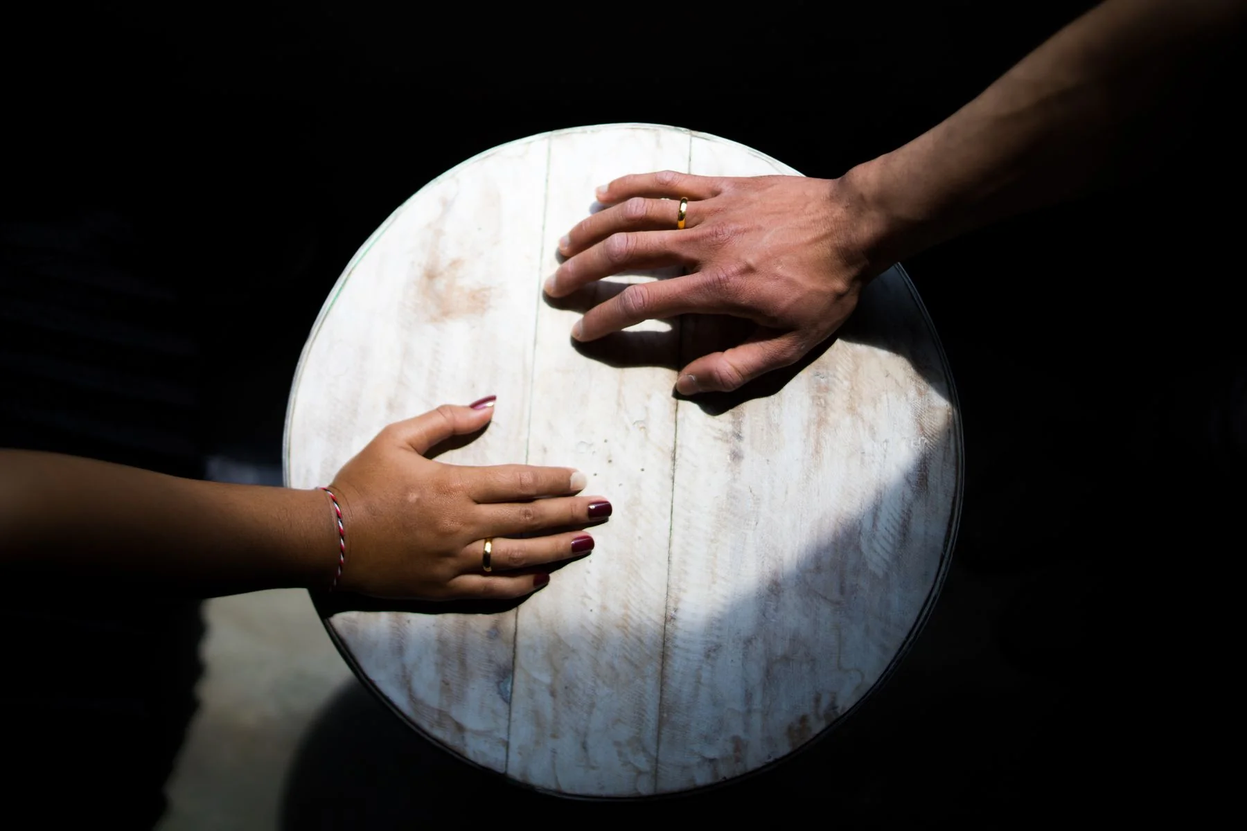 Two hands wearing plain wedding bands rest on opposite sides of a round wooden table.