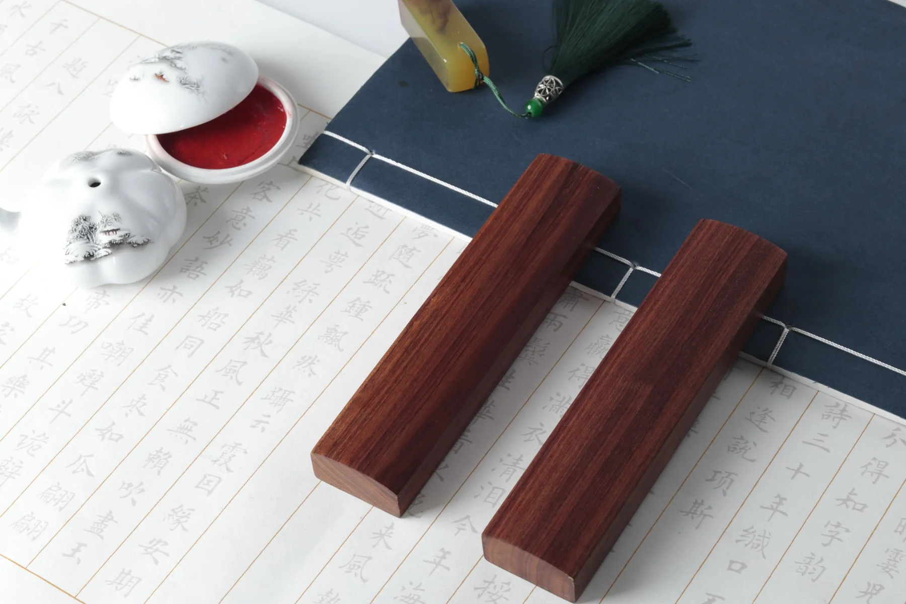 Chinese calligraphy paper, wooden paperweights and a dark cloth-bound book beside a porcelain seal-paste dish.